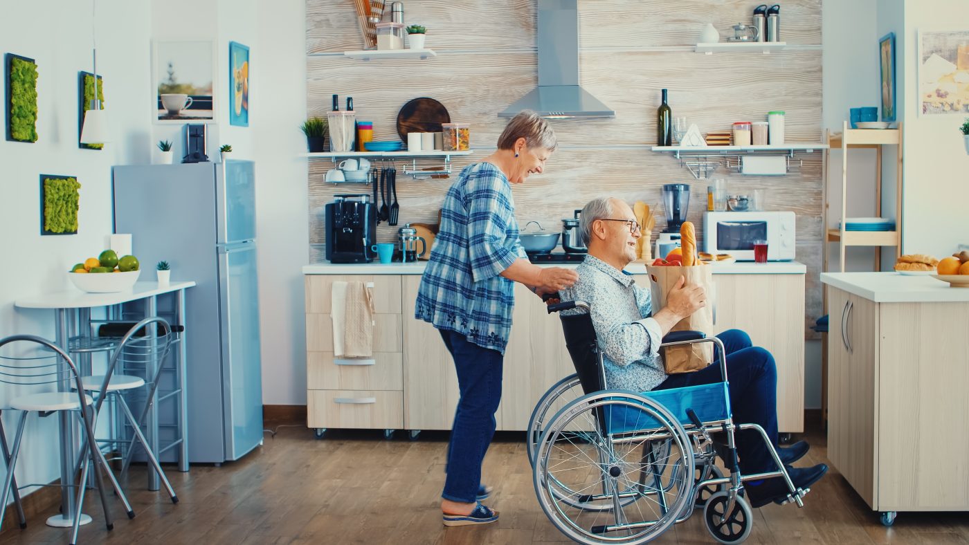 old woman pushing invalid senior husband wheelchair after arriving with grocery paper bag from supermarket mature people with fresh vegetables cooking breakfast -